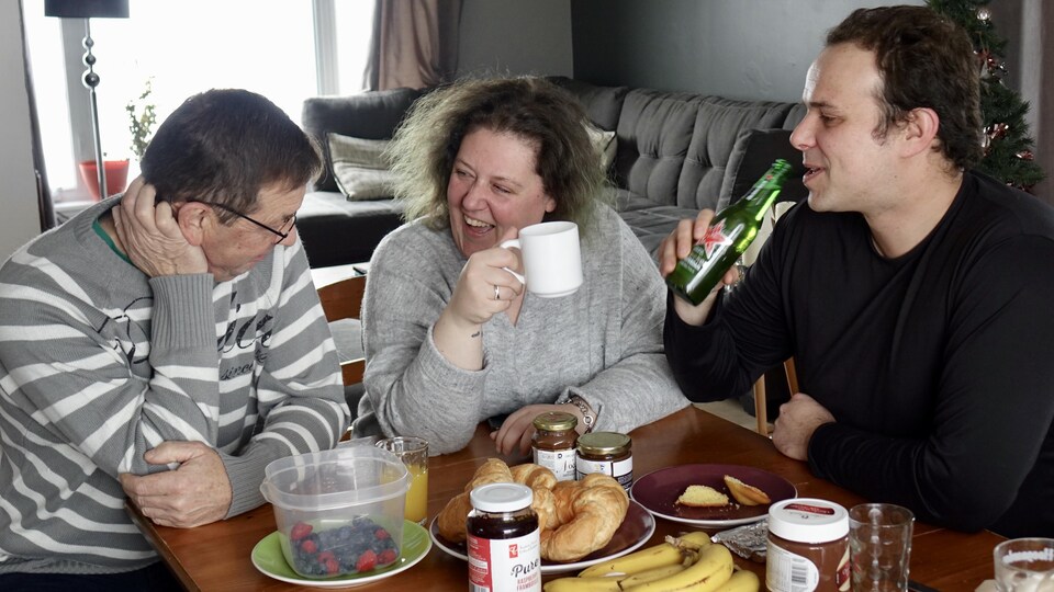 Trois personnes sont assises autour d'une table en bois et dégustent un déjeuner. Sur la table il y a des bananes, des croissants, des petits fruits et du chocolat.