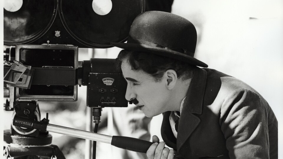 Un homme avec un chapeau melon et une moustache, le dos recourbé, en train de manœuvrer une vieille caméra. 