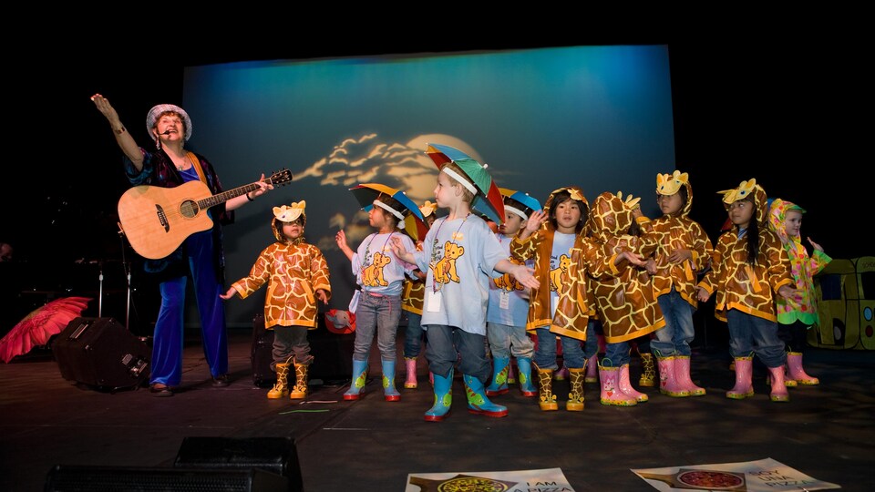 Charlotte Diamond avec une guitare et un groupe d'enfants qui portent des imperméables sur une scène.