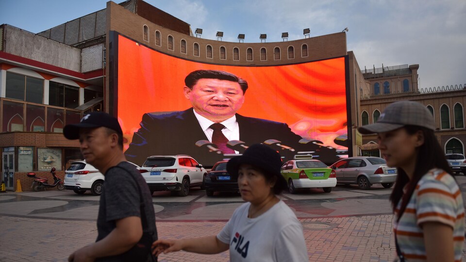 Un homme et deux femmes marchent devant un immeuble où est projetée l'image du président Xi Jinping.