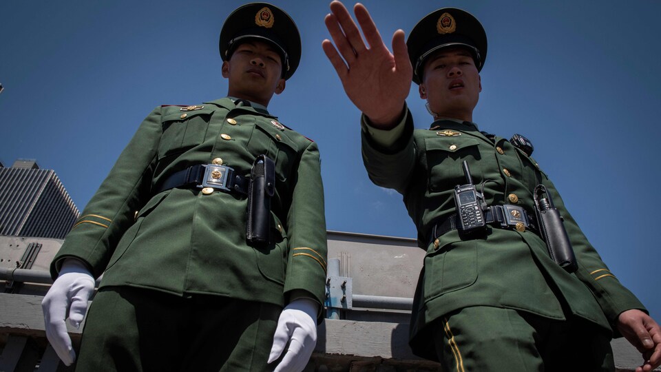 Deux policiers chinois en uniforme. L'un d'eux lève la main pour repousser un photographe.