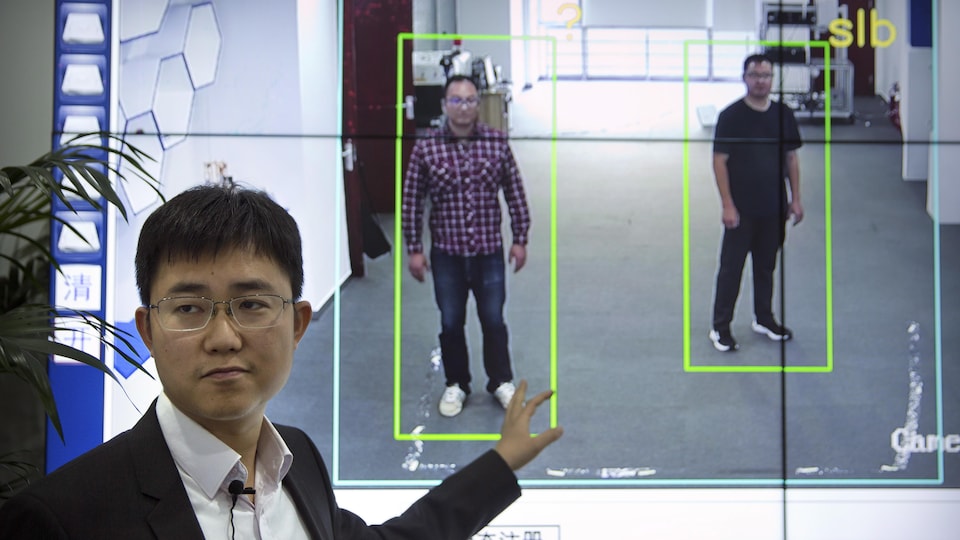 Un homme en complet pointe un écran où on peut apercevoir l'image de deux hommes, debout, apparaissant dans des cadres verts.