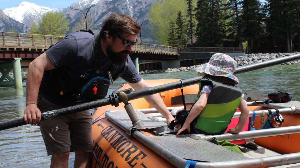 Chris Mc Kenna , debout à côté d'un bateau pneumatique où se trouve un enfant, sur une rivière.