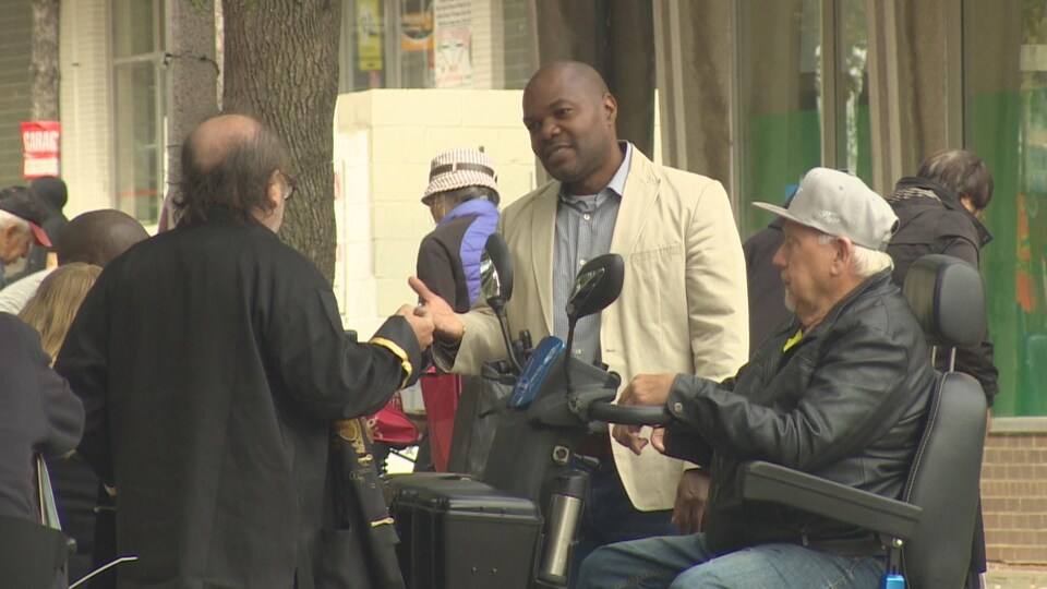 Chris Moise parlant avec plusieurs citoyens, devant une épicerie.