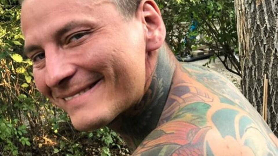 Un homme tourne la tête par dessus son épaule pour sourire à l'objectif. Son cou et son dos sont recouverts de tatouages. Derrière lui se trouvent des buissons et un arbre.