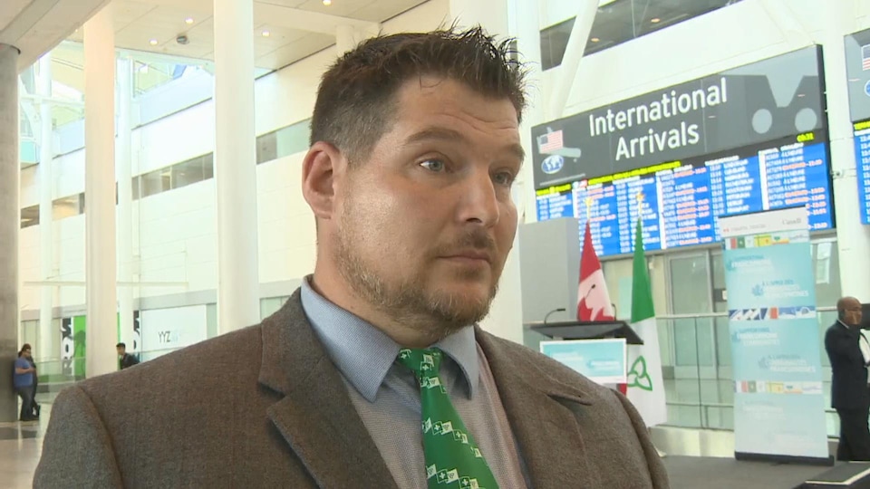 Photo d'un homme en complet portant une cravate avec des drapeaux franco-ontariens devant le tableau d'affichage des arrivées à l'aéroport