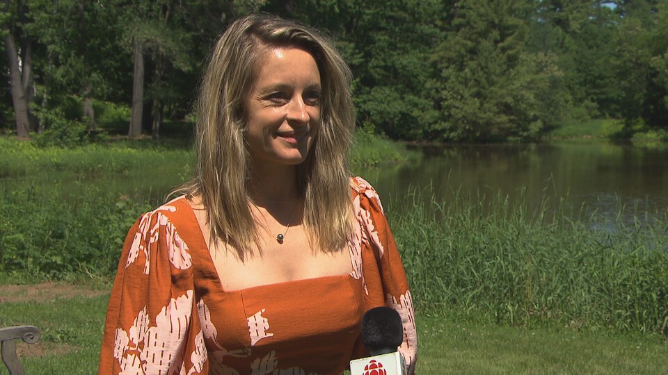 Une dame répond aux questions devant un micro de Radio-Canada. Des arbres et un lac se situent derrière elle.
