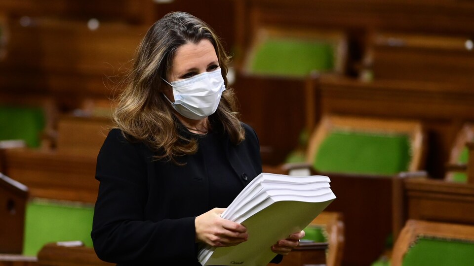 Chrystia Freeland, portant un masque et se tenant debout à la Chambre des communes, tient une pile de documents.
