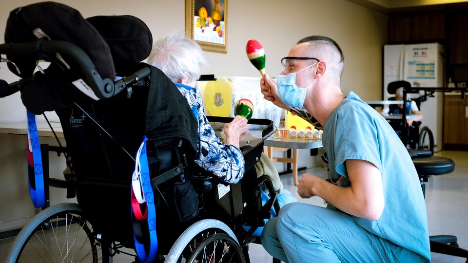 Un préposé masqué tient un maracas à côté d'une dame en fauteuil roulant vue de dos. Elle aussi tient un maracas.