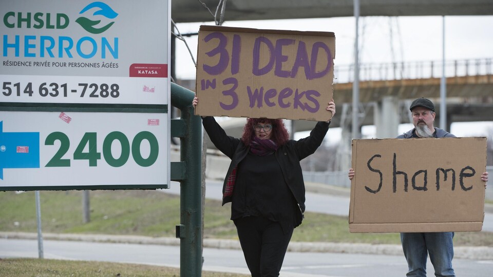 Une femme et un homme protestent devant le CHSLD Herron. La femme porte une pancarte où est écrit en anglais : « 31 morts en 3 semaines », alors que la pancarte de l'homme dit : « honte ».