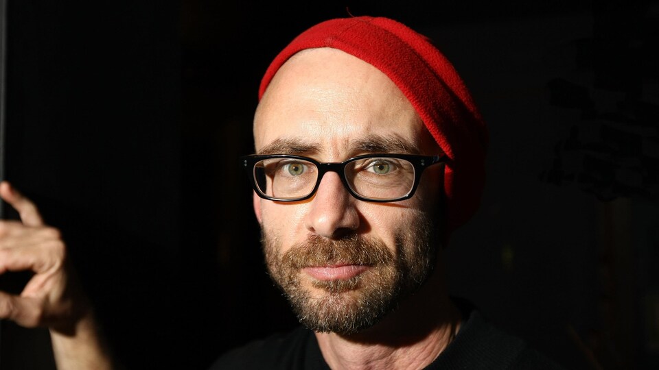Un homme portant des lunettes et une tuque rouge fixe l'objectif de l'appareil photo. 