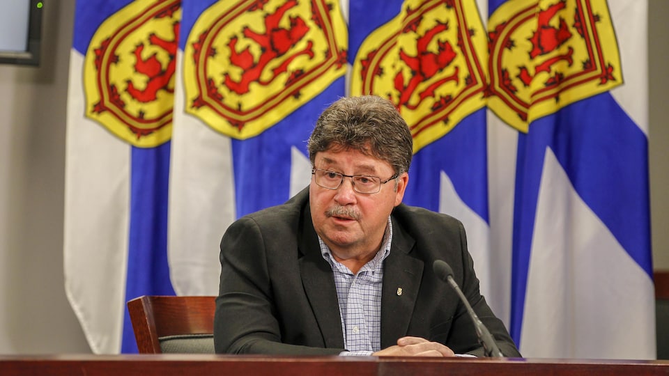 Chuck Porter en conférence de presse, assis seul à une table devant des drapeaux de la Nouvelle-Écosse.