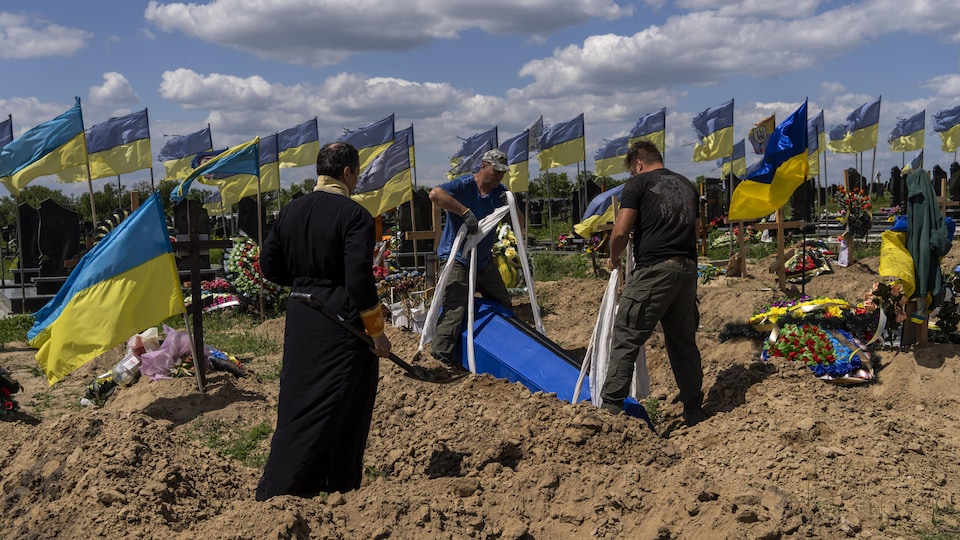 Dos hombres llevan un ataúd azul en el suelo, bajo la mirada de un sacerdote ortodoxo.  Muchas banderas ucranianas ondean al viento. 
