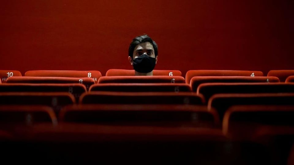 Un homme, portant un masque noir, est assis dans une salle de cinéma. 