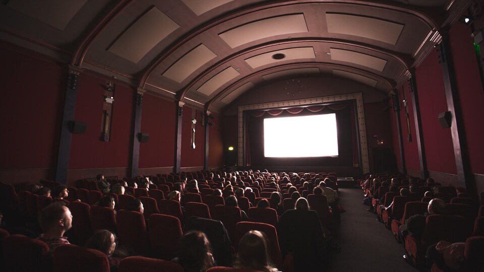 Une salle de cinéma éclairée par le grand écran. Quelques personnes sont assises dans la salle.