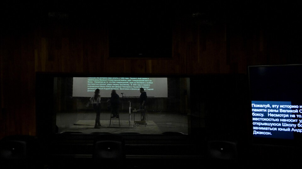 Trois personnes assises devant un écran sur lequel est affiché un texte défilant, dans un studio de doublage.