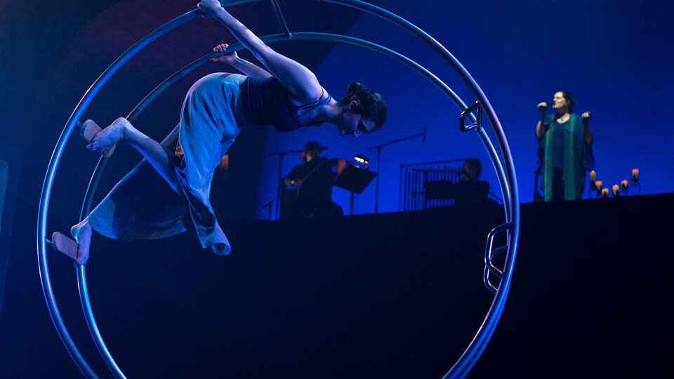 Une acrobate se tient dans une roue de cirque. Dans la pénombre en arrière-plan, il y a plusieurs musiciens. 