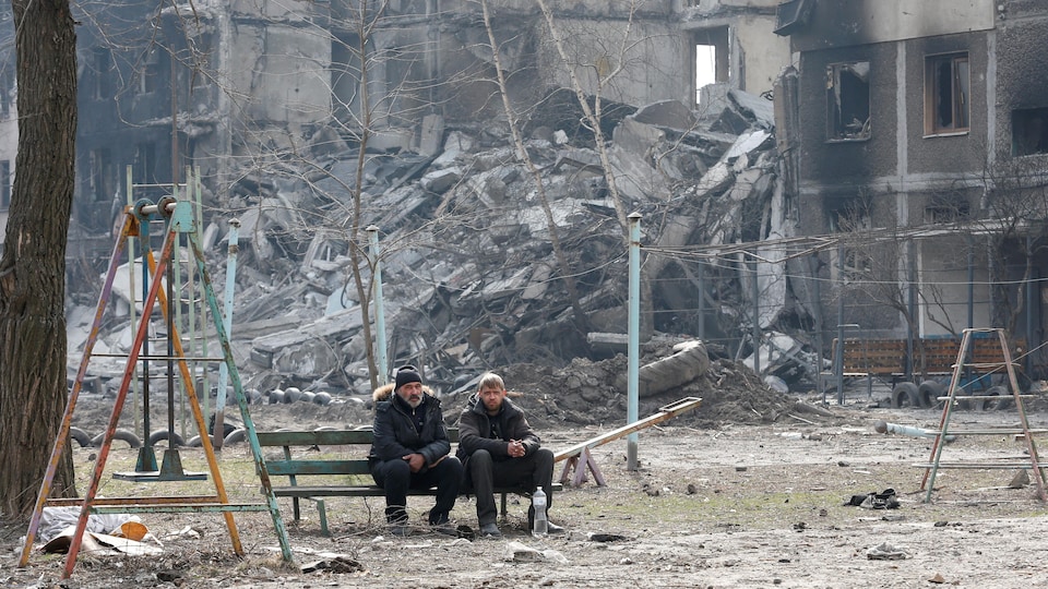 Deux hommes assis sur un banc de parc près d'un grand immeuble en ruine.
