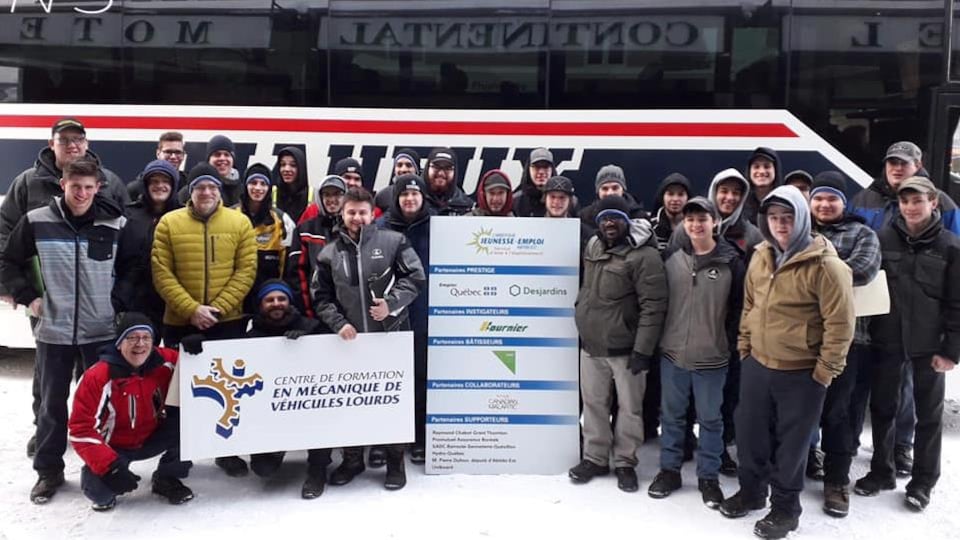 De jeunes adultes sont réunis autour d'une affiche du Centre de formation en mécanique de véhicules lourds et du Carrefour jeunesse emploi d'Abitibi-Est, devant un autobus Maheux.