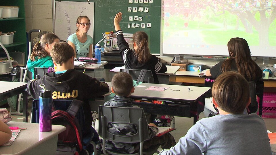 Des élèves dans une classe, dont une lève la main pour s'adresser à l'enseignante en avant.