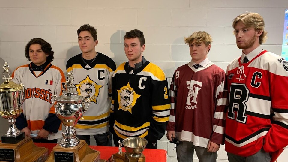 Cinq adolescents portant des chandails de hockey posent l'air très sérieux devant une table remplie de trophées.