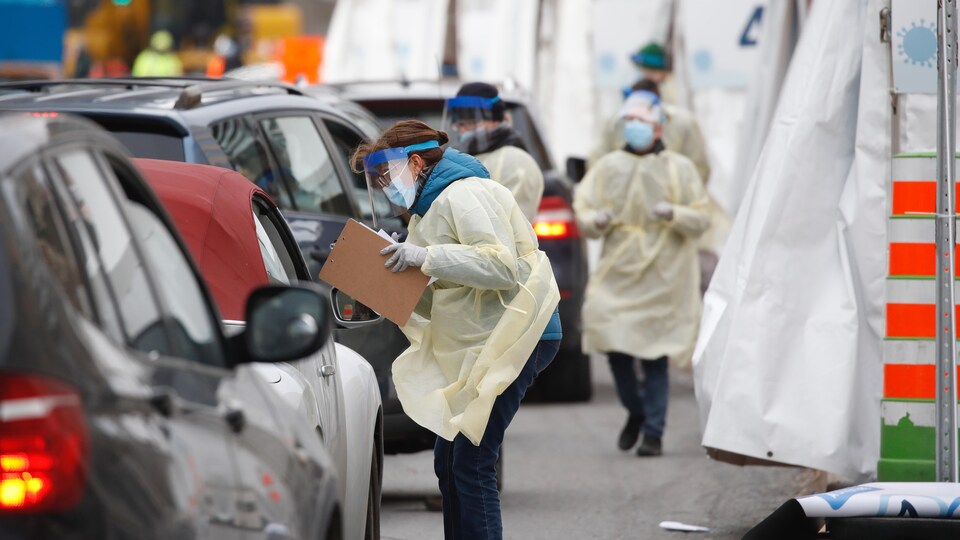 Des travailleuses de la santé portant un masque, des gants et une visière, vont à la rencontre d'automobilistes à l'extérieur de la clinique de dépistage.
