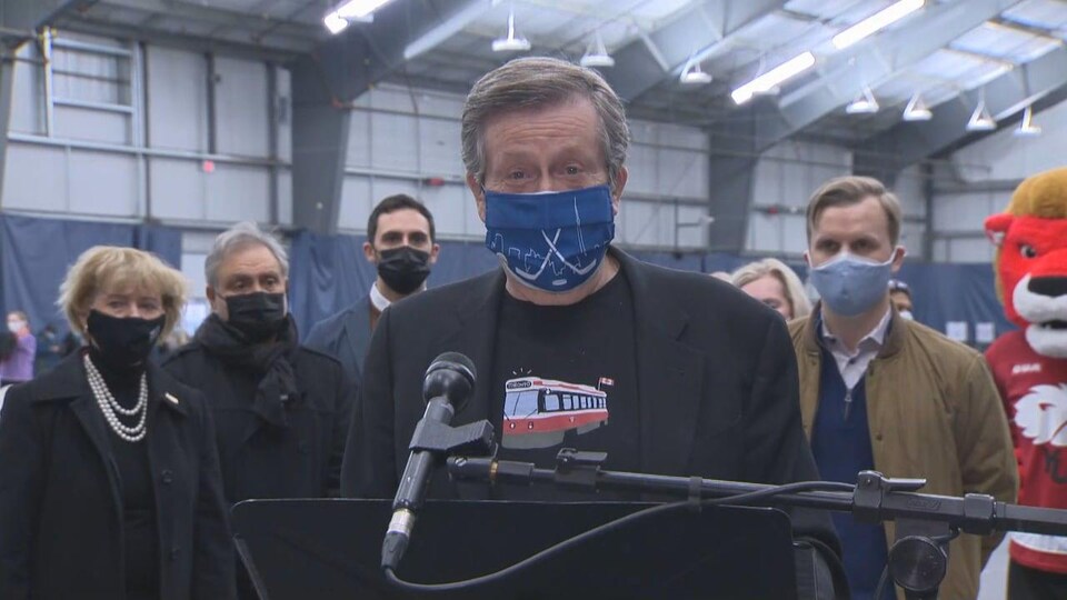 Un homme masques parle devant un micro. D'autres personnes également masquées sont derrière lui.