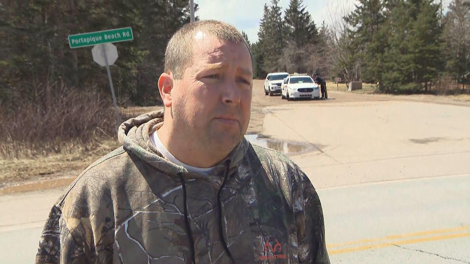 Un homme en bordure de la route, près d'un boisé, avec deux voitures de police au loin derrière lui.