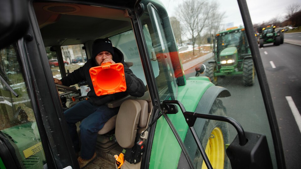 Un agriculteur au volant de son tracteur tient un cône orange en guise de porte-voix.