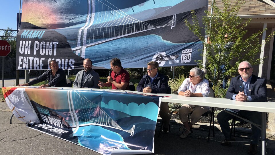 Six personnes sont assises à l'extérieur devant une grande banderole faisant la promotion d'un pont sur le Saguenay.
