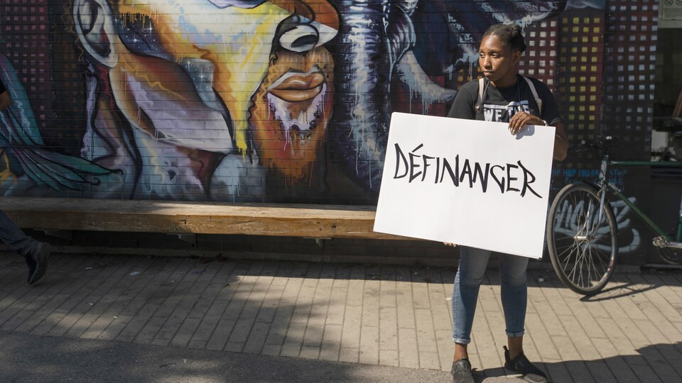 Une jeune femme tient une pancarte sur laquelle il est écrit « Définancer», devant une murale sur laquelle est peint un graffiti.