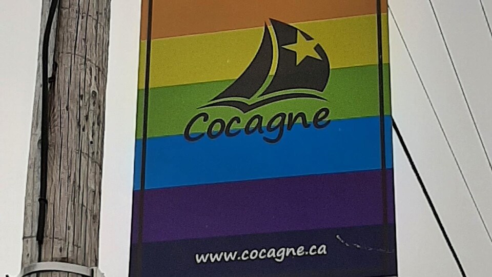 Un panneau abordant les couleurs du drapeau arc-en-ciel avec le logo de Cocagne sur un poteau d’électricité en face du bureau municipal.