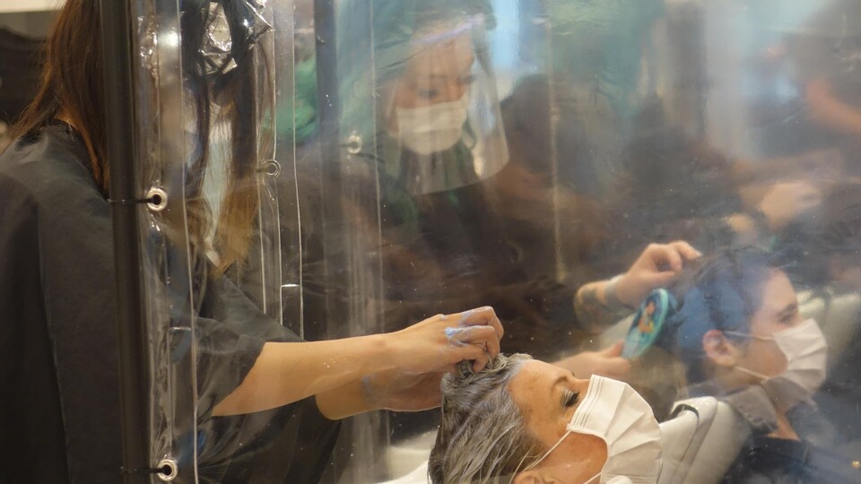 Une femme se fait laver les cheveux en portant un masque par une coiffeuse qui porte une visière