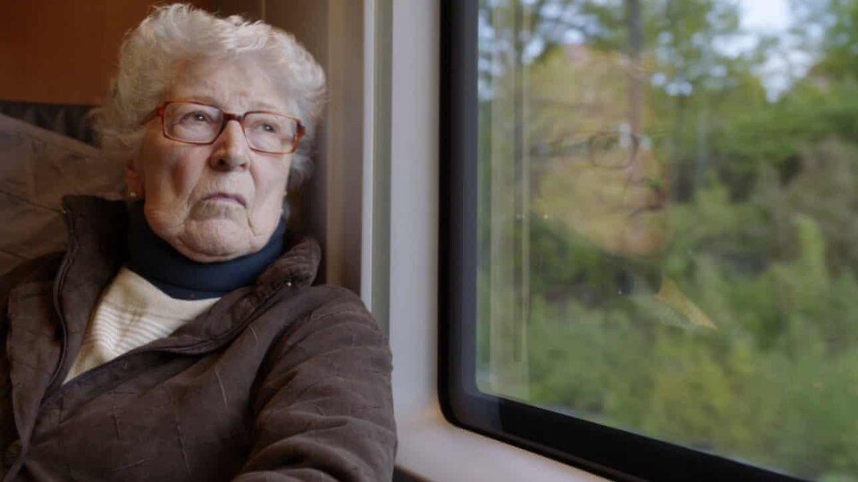 Une femme âgée regarde par la fenêtre donnant sur des arbres à l'extérieur.