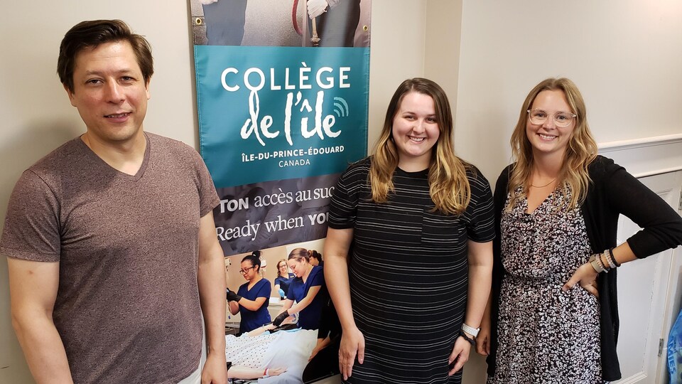 Trois étudiants posent pour la photo. Une affiche promotionnelle du collège est collé au mur derrière les étudiants.