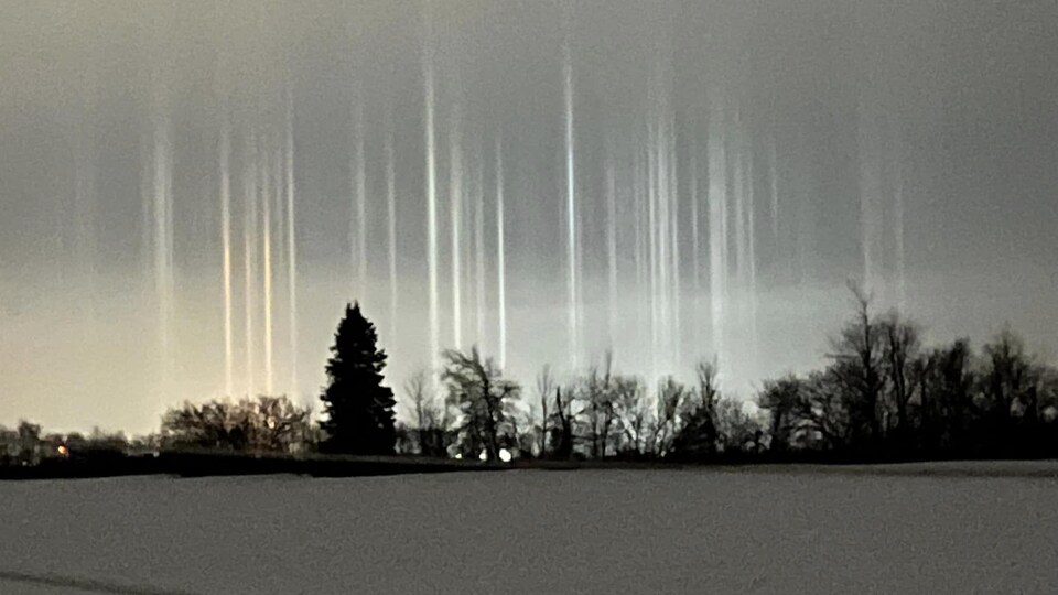 À l'horizon, au bout d'un champ en hiver, des colonnes lumineuses partent en faisceaux vers le ciel. 