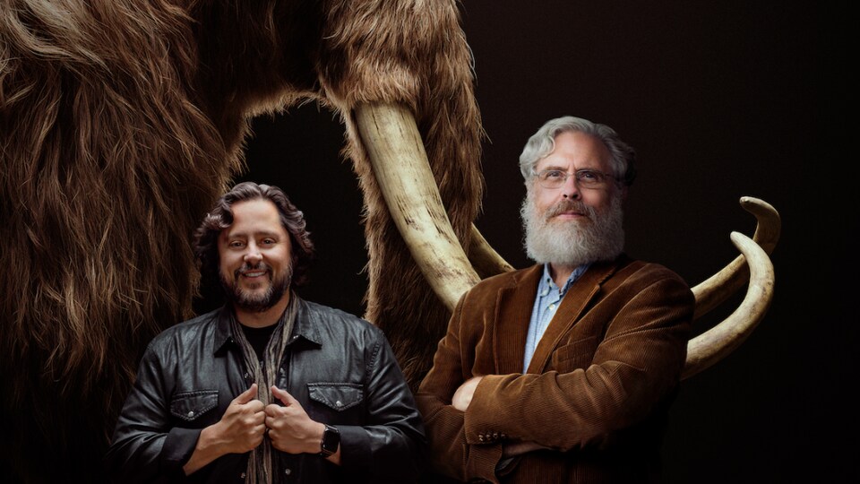 Les cofondateurs de Colossal, Ben Lamm et George Church prennent la pose devant une illustration de mammouth laineux.