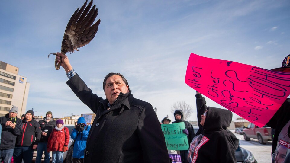 L'oncle de Colten Boushie, Alvin Baptiste, tient une aile d'aigle devant des manifestans Certains ont des pancartes autour de lui.