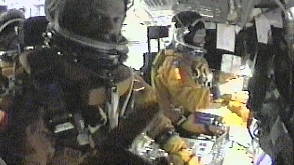 Les deux astronautes, en combinaison spatiale, s'affairent sur de l'équipement.
