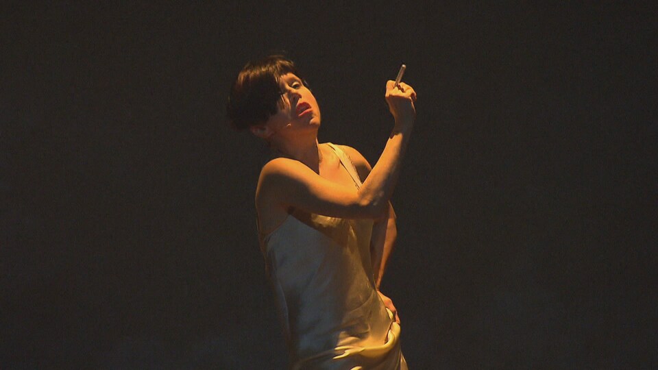 Une comédienne aux cheveux courts et vêtue d'une camisole blanche fume sur scène sous un éclairage sombre.