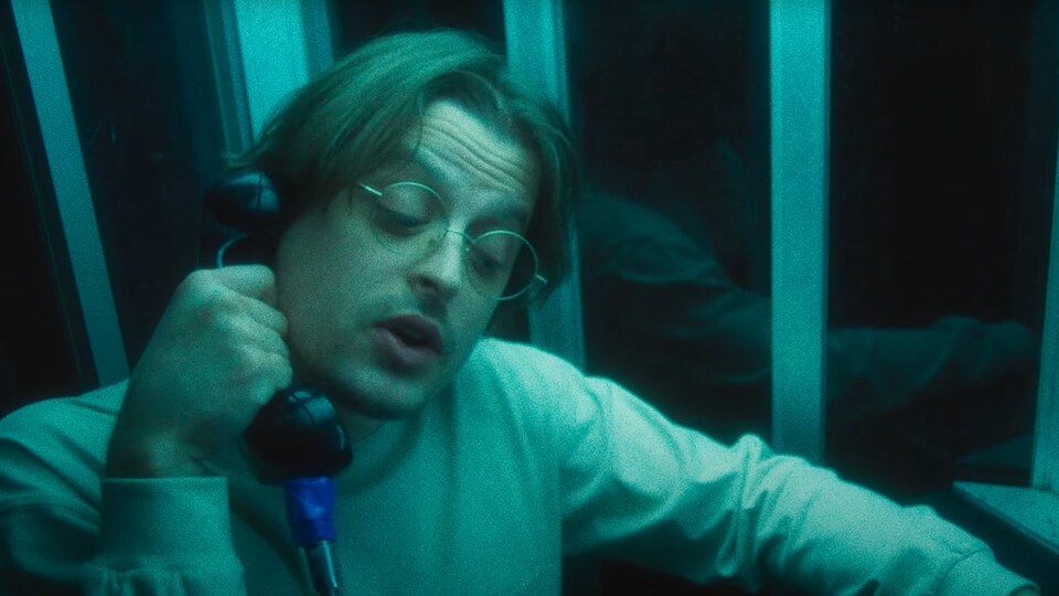 On voit le chanteur de Comment Debord parler au téléphone dans une cabine baignée de lumière bleue et verte.