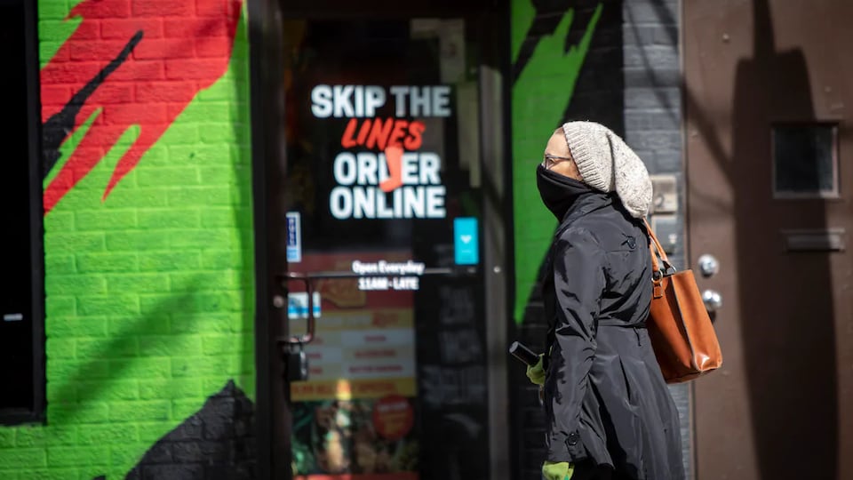 Une femme, qui se couvre le visage avec une écharpe, se tient devant la porte d'un commerce où l'on peut lire: Skip the lines, order online.