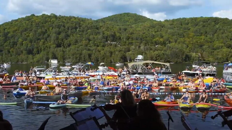 De la scène, les gens sur des kayaks, des planches, des bouées et des bateaux regardent le concert. On voit une montagne à l'arrière-plan.