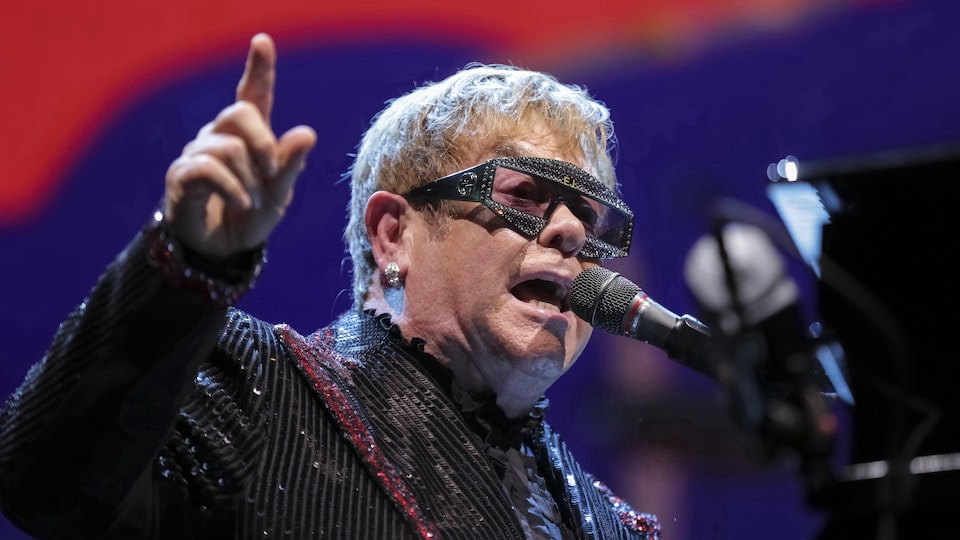 Elton John chante dans un micro, assis sur un piano, et lève sa main droite.