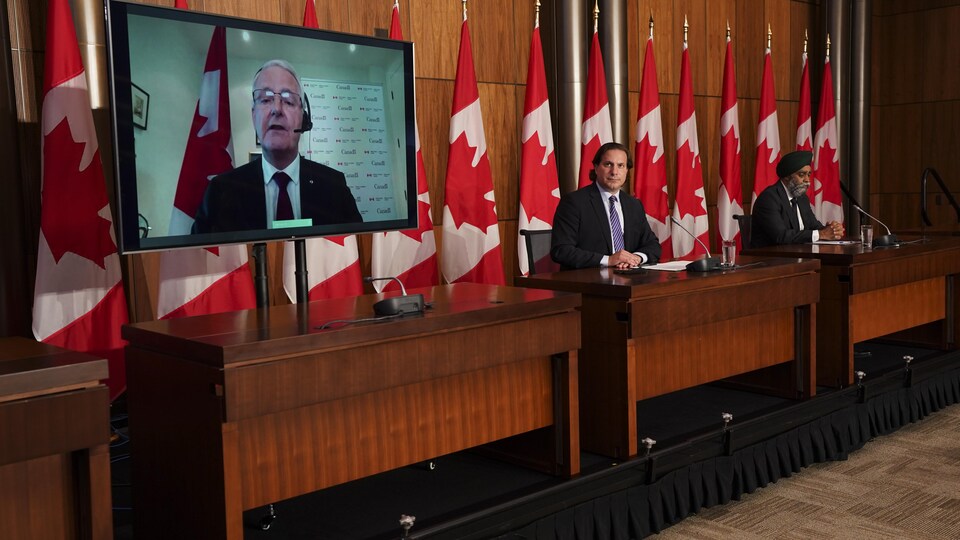 Les ministres Sajjan et Mendicino sont dans une salle de presse. Le ministre Garneau apparaît sur un écran. 