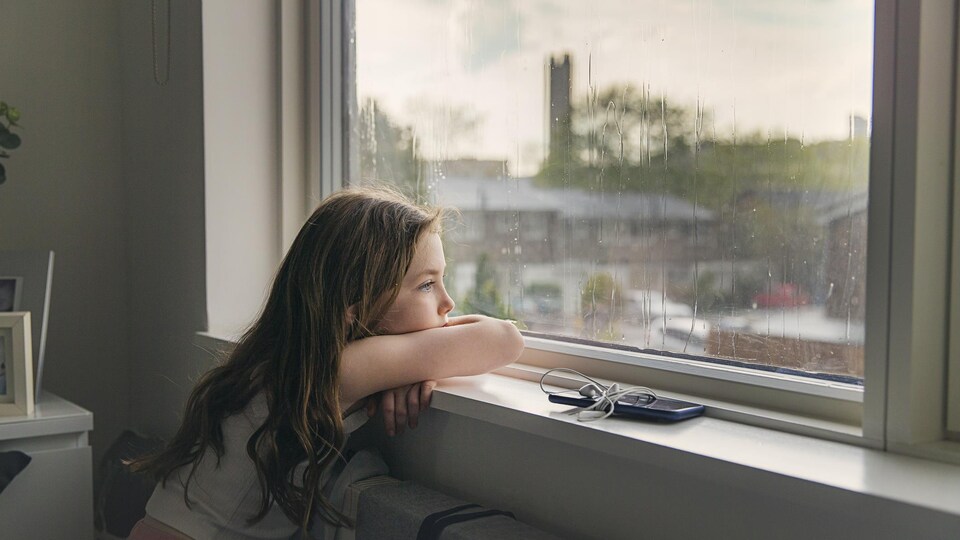 Une fillette regarde la pluie tomber, accoudée à la fenêtre.
