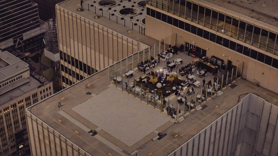 Vue aérienne d'une scène de spectacle située sur le toit d'un gratte-ciel. Des personnes soupent sur des tables autour de la scène.