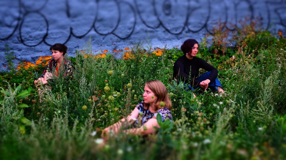 Les trois femmes sont assises dans un terrain vague rempli d'herbes et de plantes sauvages.