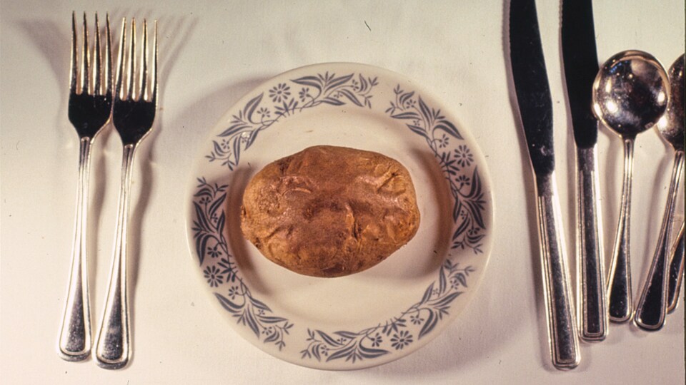 Une pomme de terre au four prend toute la place dans une assiette de faïence, comme seule vedette, sur une chic table avec nappe blanche couverts d'argent.