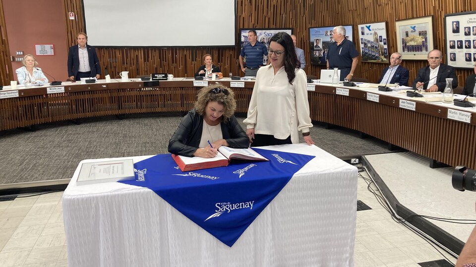 Une femme signe un livre, assise à une table, à côté de la mairesse Julie Dufour qui se tient à côté.